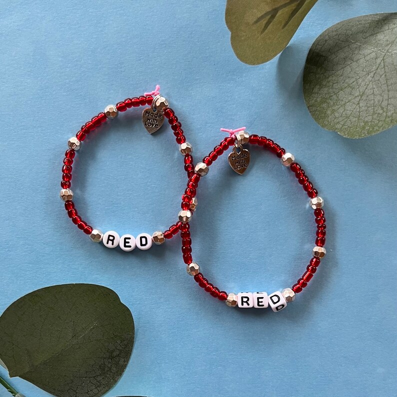 May include: Two red beaded bracelets with the word "RED" spelled out in white letter beads. The bracelets also feature silver and gold-colored beads and small heart charms that say "Made with Love".