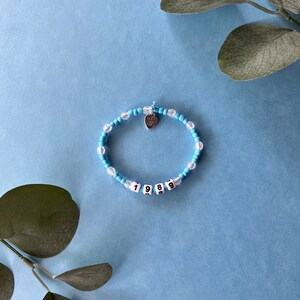 Pode incluir: Uma pulseira de contas azuis claras e transparentes. A pulseira apresenta contas brancas com a inscri&ccedil;&atilde;o "1989" e um pequeno pingente de cora&ccedil;&atilde;o prateado. A pulseira est&aacute; sobre um fundo azul claro com vegeta&ccedil;&atilde;o.