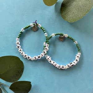 May include: Two beaded bracelets with the name "Taylor Swift" spelled out in white letter beads. The bracelets have green and clear beads, with a small heart charm that says "made with love".