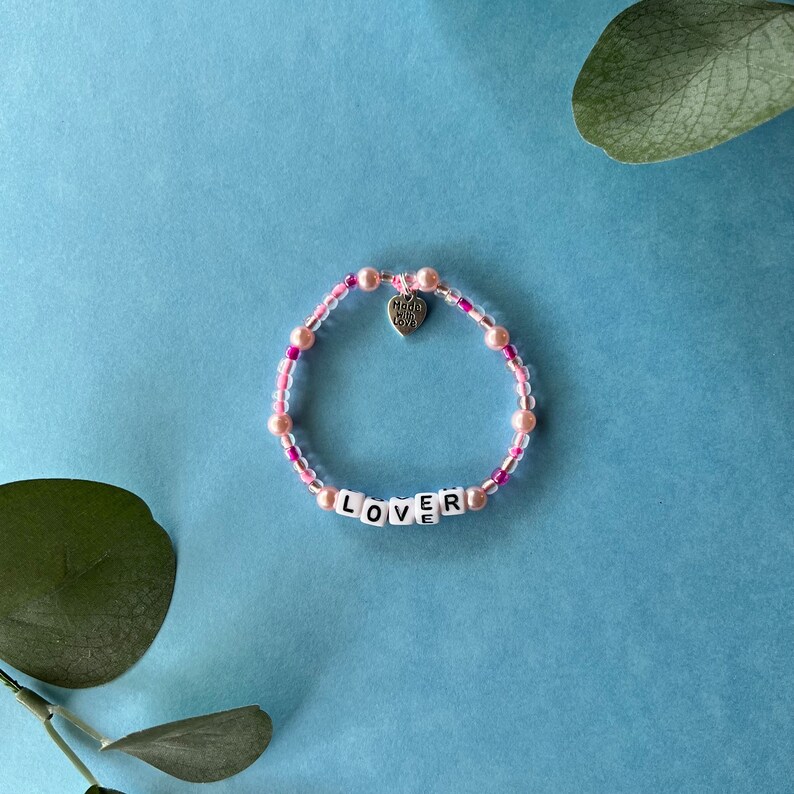 Pode incluir: Uma pulseira de contas rosa e transparentes com a palavra "LOVER" escrita com contas de letras brancas. Um pequeno pingente em forma de cora&ccedil;&atilde;o com a inscri&ccedil;&atilde;o "Made with Love" pende da pulseira. O fundo &eacute; azul claro.