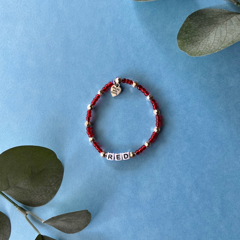 Pode incluir: Pulseira de contas vermelhas com detalhes prateados e a palavra "RED" escrita em contas brancas. Um pequeno pingente de cora&ccedil;&atilde;o com a inscri&ccedil;&atilde;o "Made with Love" pende perto do fecho. Fundo azul.