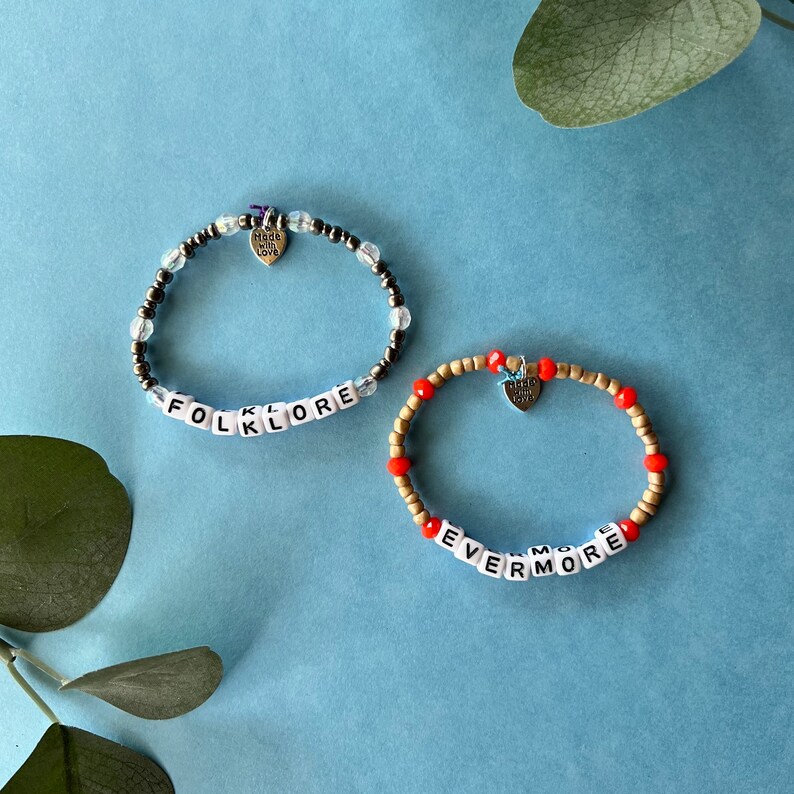 Pode incluir: Duas pulseiras de contas em uma superf&iacute;cie azul. Uma pulseira diz "FOLKLORE" com contas de letras brancas, e a outra "EVERMORE". Ambas t&ecirc;m pingentes de cora&ccedil;&atilde;o que dizem "Made with Love". As pulseiras s&atilde;o uma mistura de contas transparentes, pretas, douradas e vermelhas.