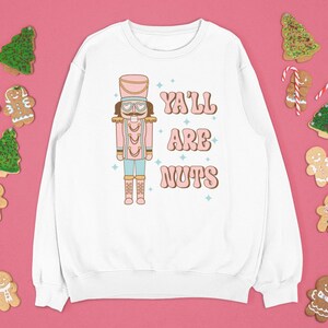 May include: White sweatshirt with a pink and blue illustration of a nutcracker and the text "Ya'll Are Nuts". The sweatshirt is surrounded by gingerbread cookies and snowflakes.