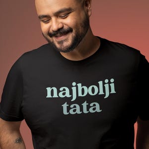 May include: Black t-shirt with the text "najbolji tata" in light blue. The shirt is worn by a smiling man with a beard and a shaved head. The background is a gradient of red and orange.