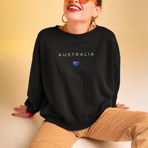 May include: A black sweatshirt with the word "AUSTRALIA" and a small Australian flag in a heart shape printed on the front. The model is wearing the sweatshirt with brown cargo pants and orange sunglasses.