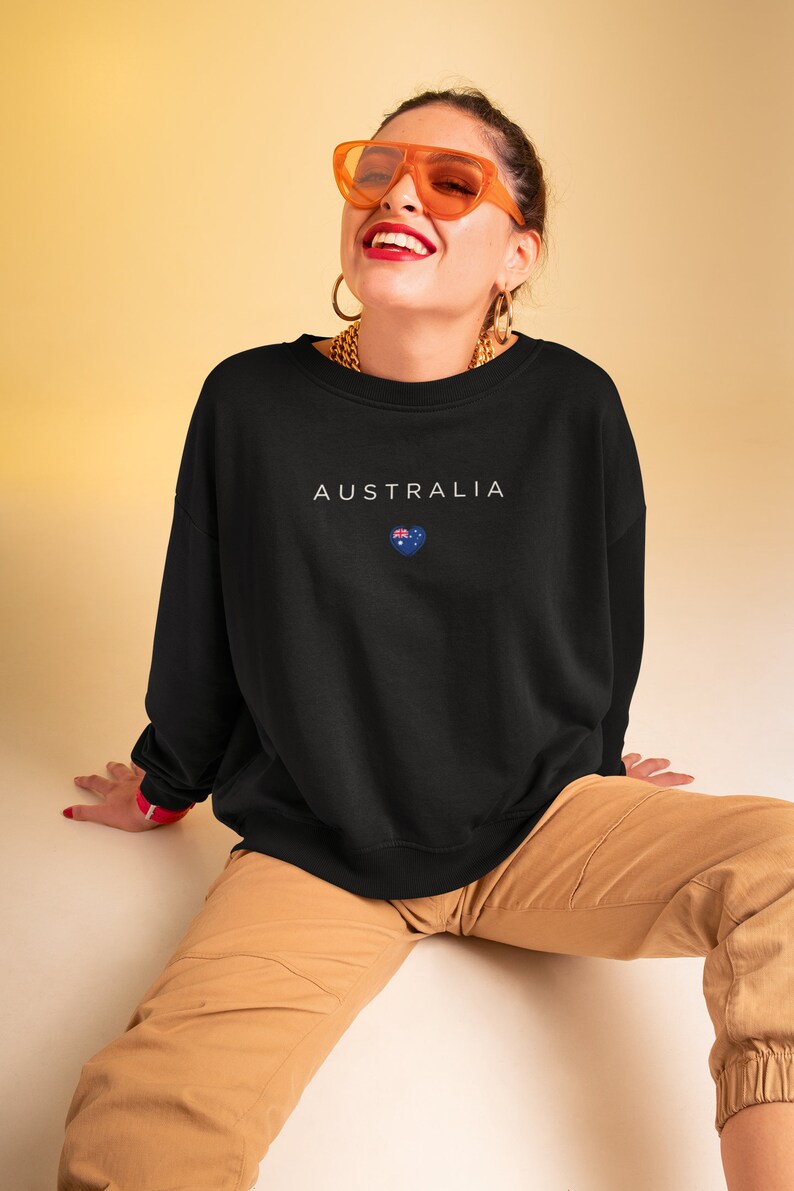 May include: A black sweatshirt with the word "AUSTRALIA" and a small Australian flag in a heart shape printed on the front. The model is wearing the sweatshirt with brown cargo pants and orange sunglasses.