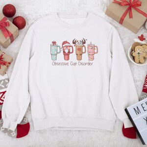 Op de afbeelding: Witte kersttrui met een afbeelding van vier mokken met kerstversiering en de tekst "Obsessive Cup Disorder".