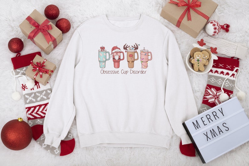 Op de afbeelding: Witte kersttrui met een afbeelding van vier mokken met kerstversiering en de tekst "Obsessive Cup Disorder".