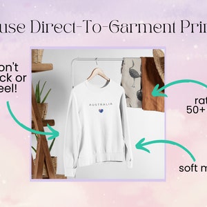 May include: White sweatshirt with the word "AUSTRALIA" and a heart with the Australian flag printed on it. The sweatshirt is hanging on a wooden rack. Text on the image reads "We use Direct-To-Garment Printing", "won't crack or peel!", "rated for 50+ washes", and "soft material".