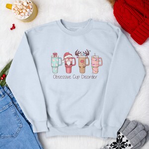 Op de afbeelding: Een lichtblauw sweatshirt met een kerstthema met vier bekers met verschillende kerstfiguren. De tekst "Obsessive Cup Disorder" staat onder de bekers.
