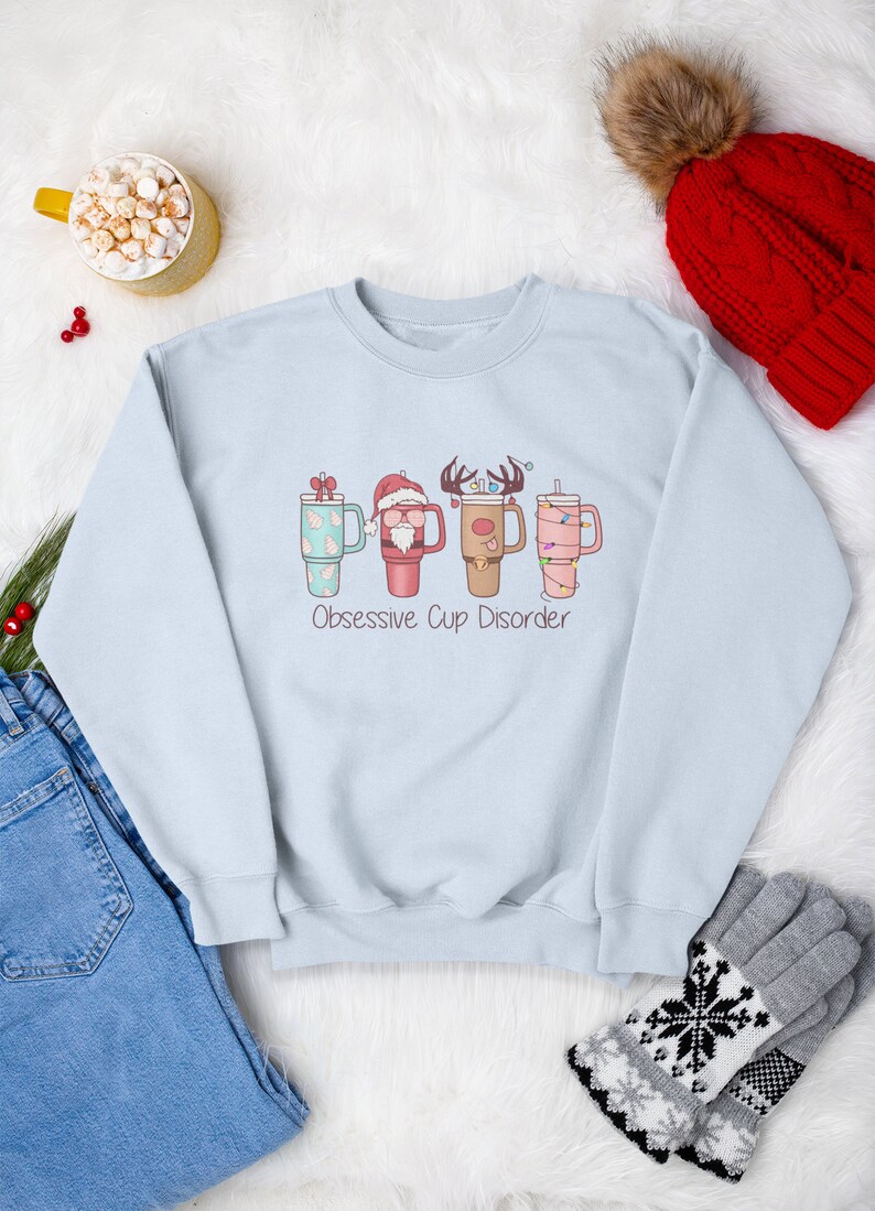 Op de afbeelding: Een lichtblauw sweatshirt met een kerstthema met vier bekers met verschillende kerstfiguren. De tekst "Obsessive Cup Disorder" staat onder de bekers.