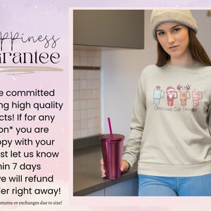 Op de afbeelding: Een lichtgrijs sweatshirt met een kerstthema ontwerp van vier mokken met verschillende dranken en de tekst "Obsessive Cup Disorder". Het model draagt ​​het sweatshirt en een grijze beanie.