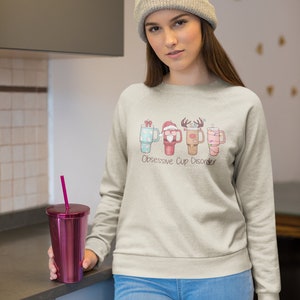 Op de afbeelding: Een licht beige sweatshirt met een afbeelding van vier kerstthemabekers en de tekst "Obsessive Cup Disorder". Het model draagt ​​het sweatshirt en houdt een roze beker met een rietje vast.