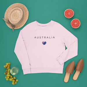 May include: A pink sweatshirt with the word "AUSTRALIA" printed on the front in black text. A small heart with the Australian flag is printed below the text. The sweatshirt is laid flat on a green surface with a straw hat, grapes, a glass of water, two grapefruit halves, and two brown leather mules.