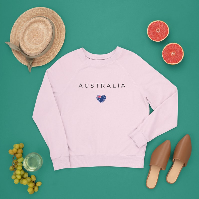 May include: A pink sweatshirt with the word "AUSTRALIA" printed on the front in black text. A small heart with the Australian flag is printed below the text. The sweatshirt is laid flat on a green surface with a straw hat, grapes, a glass of water, two grapefruit halves, and two brown leather mules.