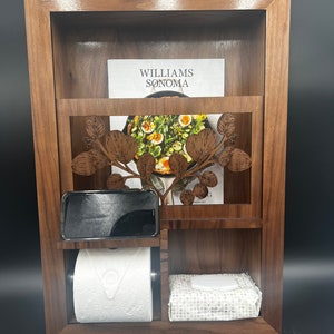 May include: A wooden wall-mounted organizer with a shelf, a phone holder, a toilet paper holder, and a tissue box holder. The organizer has a decorative cutout of a leafy branch. The shelf has a Williams Sonoma magazine on it.