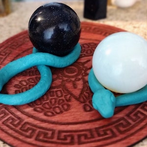 May include: Two teal-colored snake figurines coiled around spherical objects. One snake holds a dark blue, glittery sphere, while the other snake holds a smooth, opaque white sphere. Both snakes are positioned on a circular, reddish-brown wooden base.