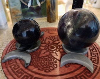 May include: Two polished stone spheres resting on gray crescent-shaped stands. The left sphere is dark blue with gold glitter flecks, while the right sphere is a deep purple with swirling patterns. Both sit on a round wooden base with etched designs.