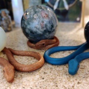 May include: Three spherical stones resting on coiled snake-shaped stands. One stone is opaque white, one is mottled green and gray, and one is dark brown. Two stands are textured brown, and one stand is a smooth, matte blue color.