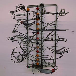 May include: A wall-mounted kinetic marble run sculpture constructed from dark grey metal wire. The structure features a central vertical column with alternating red and orange spheres, surrounded by complex wire tracks, loops, and funnels.
