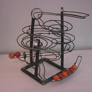 May include: A kinetic sculpture featuring a dark metal wire frame with multiple spiraling tracks and a square base. Several small, smooth spheres in shades of orange and red are positioned along the lower tracks and the base of the structure.