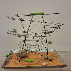 May include: A kinetic sculpture with a complex wire frame structure. The sculpture features multiple levels and loops, with green marbles rolling through the pathways. The base contains sand and small rocks. The overall design is intricate and visually engaging.