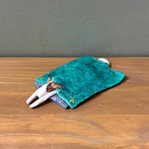 May include: A small clay figurine of a dog with brown and white fur lying on a gray bed, covered by a textured teal blanket. The blanket features a small yellow paw print detail near the corner, and the dog's head and front paws are visible.