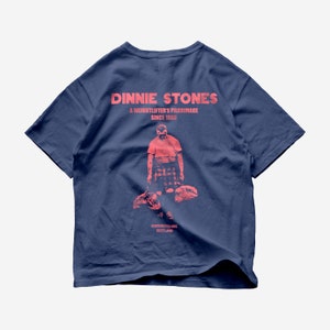 May include: A navy blue t-shirt with a pink graphic print of a man in a kilt lifting stones. The text "Dinnie Stones, A Weightlifter's Pilgrimage Since 1900, Aberdeenshire Scotland" is printed on the back of the shirt.