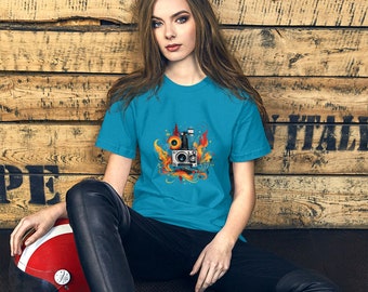 Op de afbeelding: Een turquoise blauw t-shirt met een afbeelding van een vintage stereosysteem met luidsprekers en een platenspeler. De afbeelding is omgeven door oranje en gele vlammen en swirls.