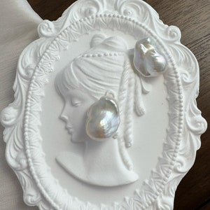 Puede incluir: Dos perlas barrocas iridiscentes descansan sobre una placa blanca ornamentada estilo camafeo. La placa presenta el perfil de una mujer y está rodeada por un borde decorativo. Las perlas tienen un brillo lustroso, reflejando la luz. El fondo es una superficie de madera oscura.