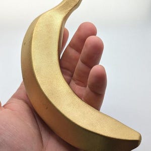 Könnte beinhalten: Eine goldene Bananenskulptur mit strukturierter Oberfläche. Die Banane ist gebogen und wird in einer Hand vor weißem Hintergrund gehalten. Die Skulptur hat ein mattes Finish und ist ein dekoratives Objekt.