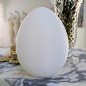 May include: A large, smooth, white egg-shaped object. The egg is the focal point, with a blurred background including a decorative glass vase and a marble surface. The egg's shape is prominent, suggesting a decorative or artistic piece.