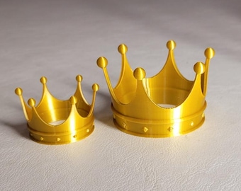 May include: Two gold-colored crowns, one smaller than the other, are displayed on a neutral surface. The crowns have a classic design with rounded points and a circular base. The crowns are made of a shiny material.