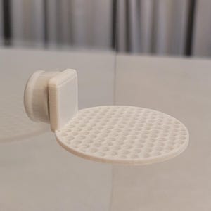 May include: A white, 3D-printed wall-mounted shelf. The shelf has a circular platform with a textured, honeycomb-like surface. It features a rectangular back piece and a cylindrical mounting component.