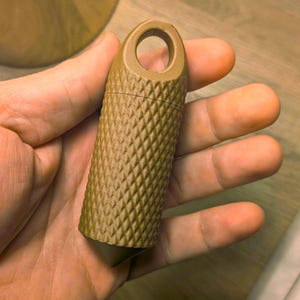 Puede incluir: Un recipiente cilíndrico de color verde oliva con un patrón de diamantes y un lazo en la parte superior. La parte inferior del recipiente es puntiaguda. El recipiente se sostiene en una mano.