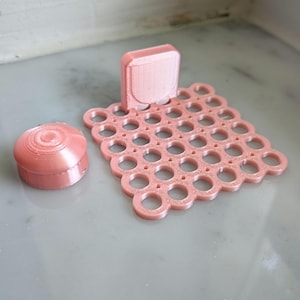 May include: A light pink 3D printed object consisting of a square grid base with circular cutouts and a small vertical tab, accompanied by a separate small, round, domed cap with concentric circular ridges on its top surface.