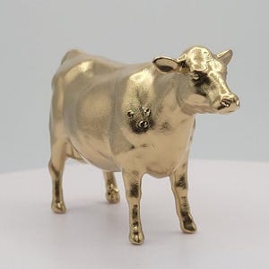 Op de afbeelding: Een goudkleurig koeienbeeldje. De koe staat met het hoofd iets naar rechts gedraaid. Het beeldje heeft een gladde, metallic afwerking en is een decoratief item.