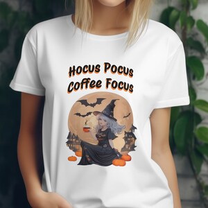 May include: White t-shirt with a graphic of a witch holding a cup of coffee in front of a full moon with bats flying around it. The text "Hocus Pocus Coffee Focus" is printed on the shirt.