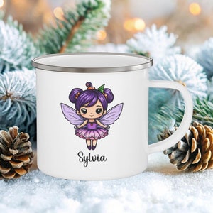 May include: White enamel mug with a silver rim and handle. The mug features a cartoon fairy with purple hair, wings, and a pink dress. The name "Sylvia" is written below the fairy. The mug is set against a snowy background with pinecones and evergreen branches.