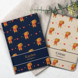 May include: Two personalized passport covers with a cute cartoon capybara design. One cover is navy blue with gold stars and hearts, and the other is beige with gold stars and hearts. The navy blue cover has the name "Kayden" and the beige cover has the name "Celeste". Both covers say "Passport" underneath the name.