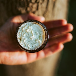 May include: A close-up of a small glass jar containing a white cream. The jar is being held in someone's hand.