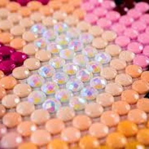 May include: Close-up of a diamond painting in progress, featuring a variety of pink, orange, and white rhinestones. The rhinestones are arranged in a geometric pattern.