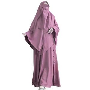 May include: A mauve-colored modest dress ensemble. The outfit includes a long dress with buttons down the front and a matching head covering with a face veil. The sleeves are long, and the fabric appears to be lightweight.