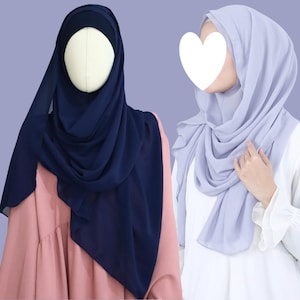 Puede incluir: Se muestran dos hiyabs. Uno es azul marino oscuro, drapeado sobre un maniquí. El otro es lavanda suave, usado por una persona. Ambos están hechos de una tela fluida y ligera, con pliegues y drapeados suaves.