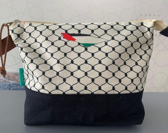 May include: A white and black patterned toiletry bag with a black bottom. The bag has a zipper closure and a brown strap. The pattern features a black mesh design with a Palestinian flag in the center.