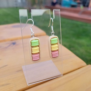 Könnte beinhalten: Ein Paar Hängeohrringe mit drei übereinander gestapelten Macarons in Grün-, Gelb- und Rosatönen. Die Macarons sind aus Polymer Clay gefertigt und an silberfarbenen Haken befestigt.