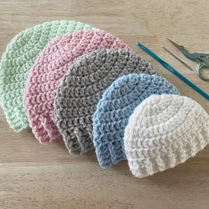 Puede incluir: Seis gorros de ganchillo hechos a mano en tonos pastel: verde menta, rosa, gris, azul claro y blanco. Un ganchillo, una aguja y unas tijeras pequeñas se colocan junto a los gorros. Los gorros están colocados sobre una superficie de madera clara.