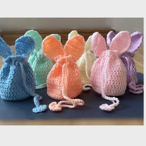 May include: Six crocheted bunny-shaped drawstring bags in various pastel colours, including light blue, pink, peach, yellow, and lavender. The bags are made with a soft, textured yarn and have long, floppy ears.