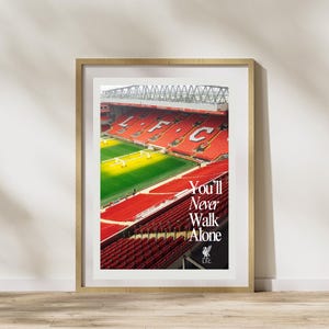 Puede incluir: Impresión enmarcada que muestra un estadio con asientos rojos y un campo verde. El texto "You'll Never Walk Alone" se muestra en blanco, con el logotipo del Liverpool FC debajo. El marco es de color marrón claro.