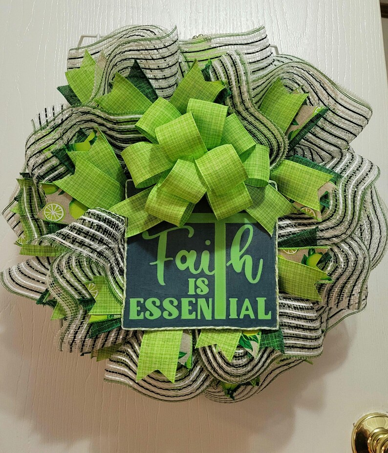 May include: A decorative wreath featuring a mix of green and black and white striped mesh and ribbons. A large green bow is centered on the wreath. A rectangular sign with the words "Faith is Essential" is attached to the wreath.